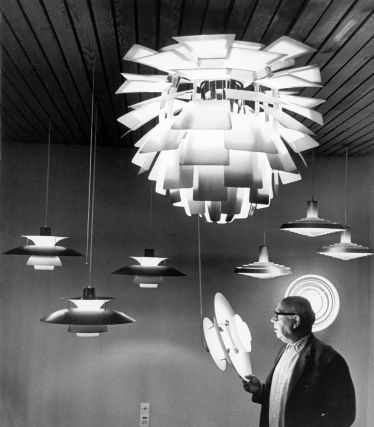 A historic black-and-white photograph of Poul Henningsen with his iconic lamps for Louis Poulsen. In the foreground, Henningsen holds a lamp model in his hand, while several of his designs hang above him—including the famous Artichoke lamp, which produces a soft, glare-free light with its many slats.