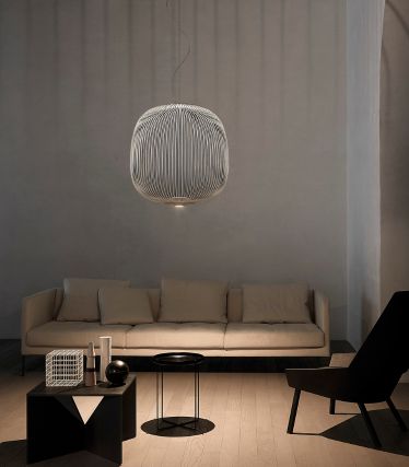 A minimalist living room with the Spokes pendant lamp by Foscarini as an elegant focal point. The lamp's delicate metal struts cast soft patterns of light on the walls and ceiling, giving the room a light, sculptural effect. A light-colored sofa, dark accents, and natural materials create a calm, modern atmosphere.